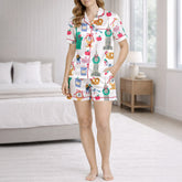 Woman wearing a white pajama set with a colorful 'Urban Wanderlust' pattern featuring city landmarks like the Statue of Liberty and Big Ben, pretzels, apples, and clocks, sitting on a white rug.