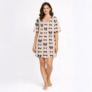 A women's short sleeve button-down pajama set with shorts, featuring a repeating pattern of small black bow ties on a light beige background. The set has black piping details and is made from a soft knit fabric.