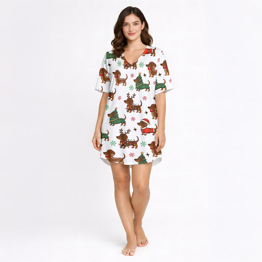 A woman wearing a white short sleeve pajama set with shorts, featuring a festive Christmas pattern of cartoon dachshunds dressed as Santa, reindeer, and Christmas trees. The pajamas have red piping.