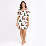 A woman wearing a white short sleeve pajama set with shorts, featuring a festive Christmas pattern of cartoon dachshunds dressed as Santa, reindeer, and Christmas trees. The pajamas have red piping.