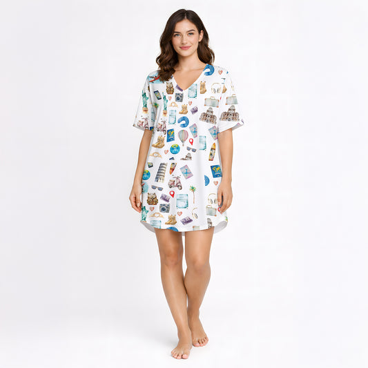 Woman wearing a white long-sleeve pajama set with a colorful watercolor travel-themed print featuring landmarks like the Statue of Liberty, Eiffel Tower, and hot air balloons, standing in a bedroom.