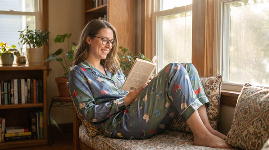 Woman wearing book-themed two-piece summer pajama set while reading in sunny window seat