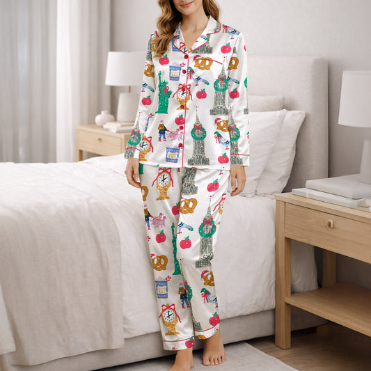 Woman wearing a white pajama set with a colorful 'Urban Wanderlust' pattern featuring city landmarks like the Statue of Liberty and Big Ben, pretzels, apples, and clocks, sitting on a white rug.