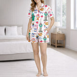 Woman wearing a white pajama set with a colorful 'Urban Wanderlust' pattern featuring city landmarks like the Statue of Liberty and Big Ben, pretzels, apples, and clocks, sitting on a white rug.