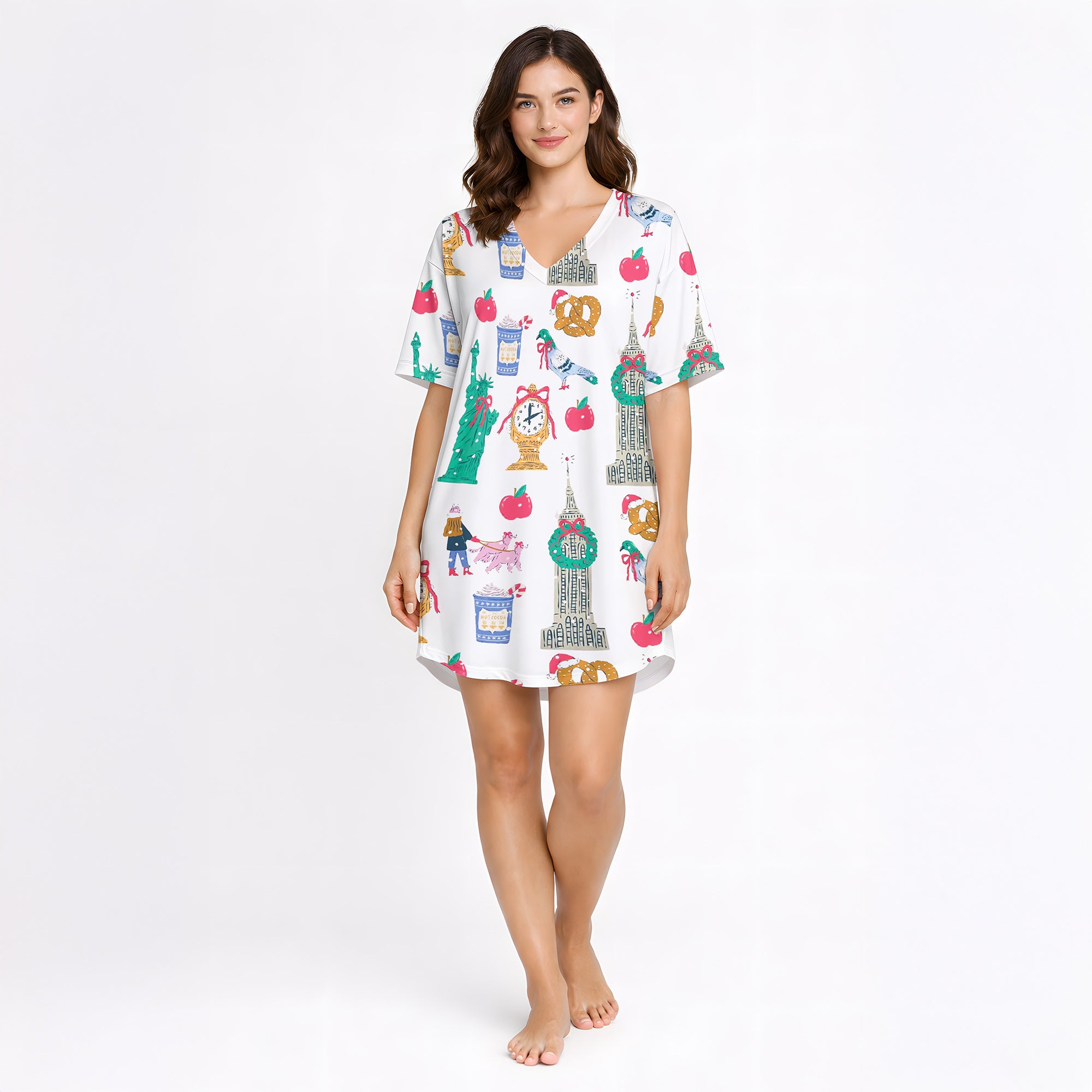 Woman wearing a white pajama set with a colorful 'Urban Wanderlust' pattern featuring city landmarks like the Statue of Liberty and Big Ben, pretzels, apples, and clocks, sitting on a white rug.