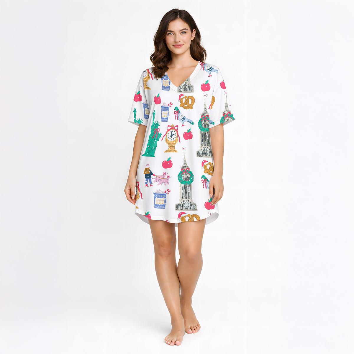 Woman wearing a white pajama set with a colorful 'Urban Wanderlust' pattern featuring city landmarks like the Statue of Liberty and Big Ben, pretzels, apples, and clocks, sitting on a white rug.